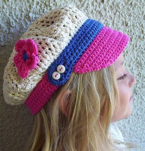 menina loira com chapéu de crochê no modelo d boné na cor cru com aplique de uma rosa, aba rosa e detalhes em azul