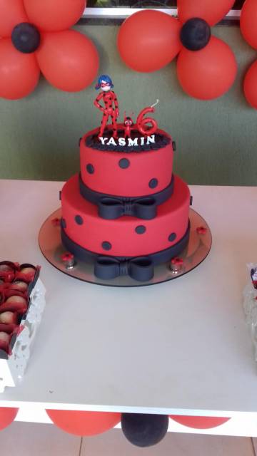 Bolo Ladybug vermelho com bolas e laços pretos e personagem, vela e nome no topo.