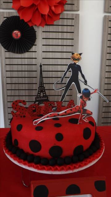 Bolo vermelho com bolinhas pretas e desenho da Ladybug, Cat Noir e da torre Eiffel em cima.