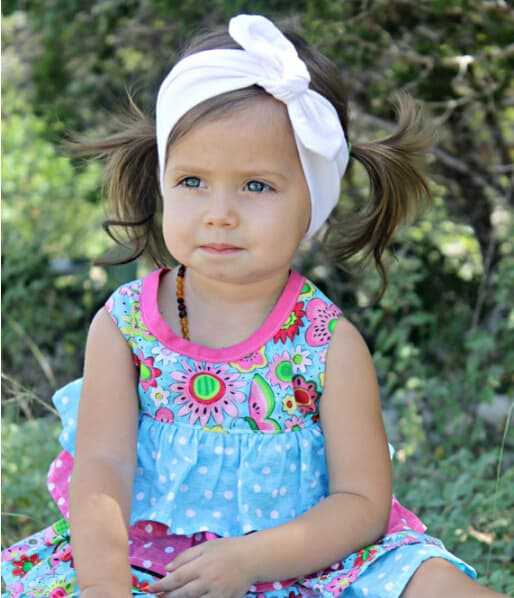 Criança usa vestido azul com rosa detalhes de estampa de flores e turbante branco ajustável de laço.