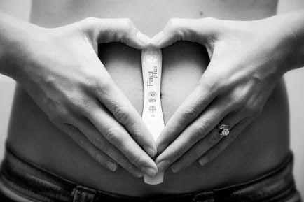 Mulher segurando teste de gravidez na frente da barriga, com as mãos formando um coração.