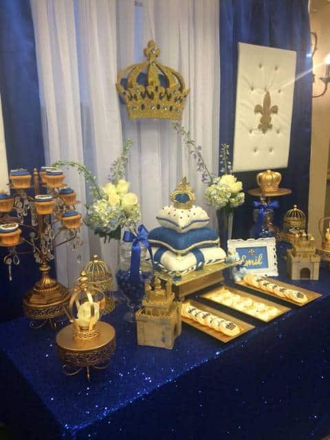Mesa e painel decorados com azul marinho, e bolo que imita uma coroa sobre almofadas.