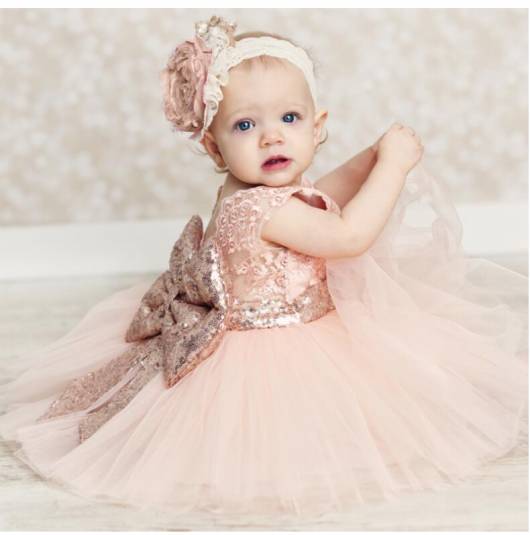 Bebê veste vestido com tule na cor rosa bege bem clarinho com laço e tiara branca com detalhes rosa na mesma cor.
