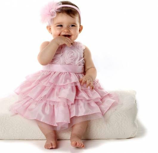 Bebê veste vestido rosa claro com babados na saia e tiara com detalhes na mesma cor.