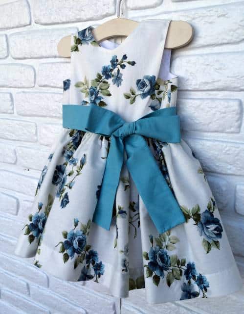 Modelo de vestido regatinha em cortegode com fundo branco e estampa floral azul com laço na mesma cor na cintura.