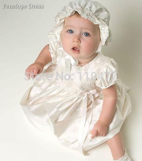 Bebê usa vestido de batizado de cetim na cor gelo com touquinha.
