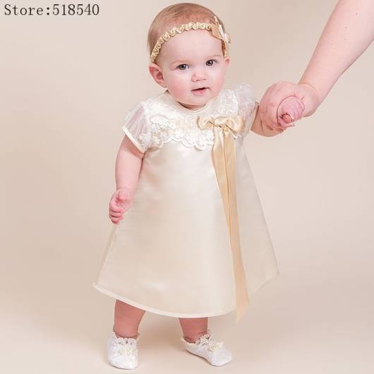 Bebê usa tiara amarelinha claro, vestido no mesmo tom de cetim em comprimento médio.