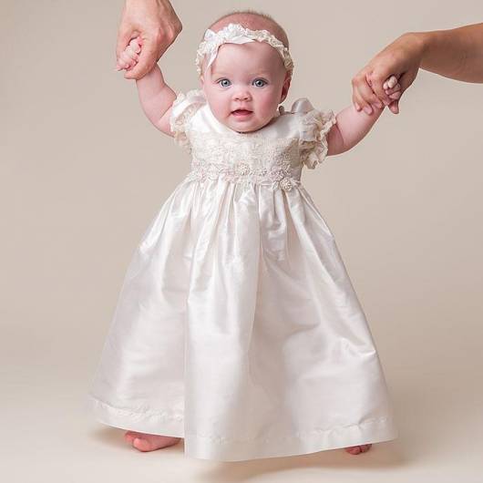Bebê usa vestido longo branco saia godê de maguinhas e tiarinha branca.
