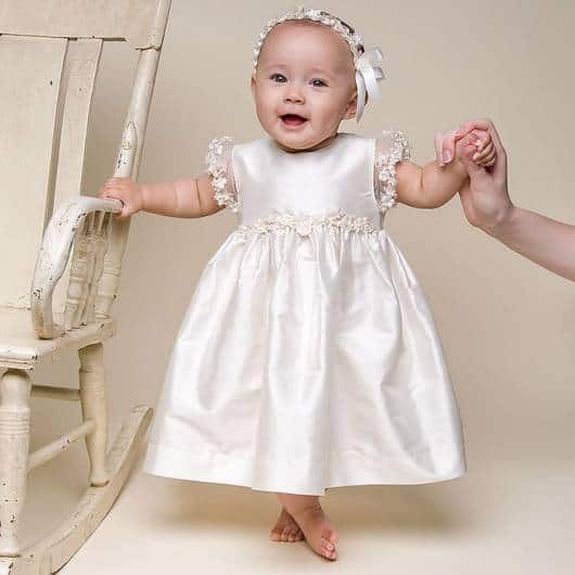 Bebê veste vestido de cetim branco comprimento médio, com tiara de tirinhas.